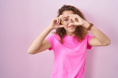 Pembe arka planda duran, elleri ve parmaklarıyla kalp şeklinde gülümseyen beyaz bir kadın. 