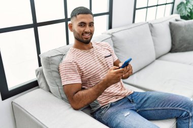 Genç Arap adam evde akıllı telefon kullanarak kendinden emin gülümsüyor.