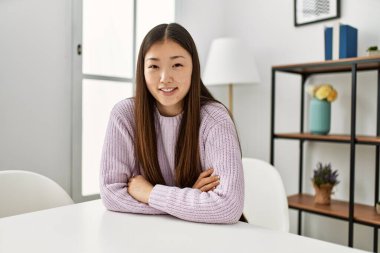 Genç Çinli kız mutlu bir şekilde masada oturuyor kollarını kavuşturup evde oturuyor..