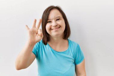 Genç Down sendromlu kadın izole edilmiş arka planda duruyor ve üç numaralı parmaklarıyla işaret ederken kendinden emin ve mutlu bir şekilde gülümsüyor.. 
