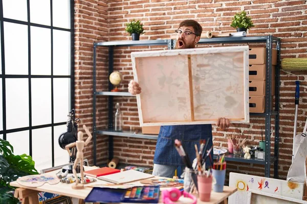 Young redhead man artist looking draw with surprise expression at art studio