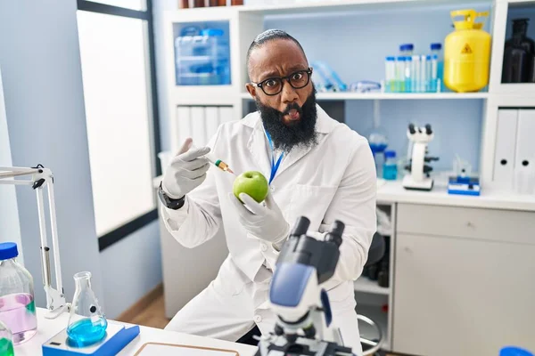 African american man working at scientist laboratory with apple making fish face with mouth and squinting eyes, crazy and comical.