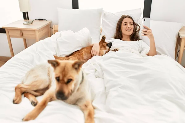 Young hispanic woman hugging dog and using smartphone lying on bed at bedroom