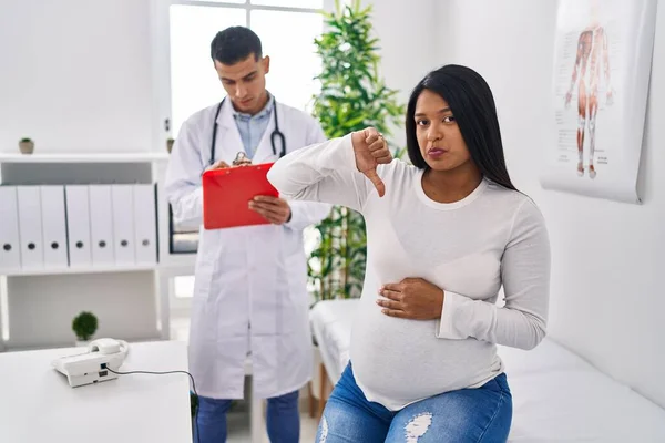 怒った顔の医者で赤ん坊を期待している若いヒスパニック系の母親 親指を下げて嫌いであることを示す負の記号 拒絶概念 — ストック写真