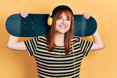 Kızıl saçlı genç bir kadın elinde kulaklık takmış mutlu ve havalı bir gülümsemeyle gülümsüyor. diş gösterme. 