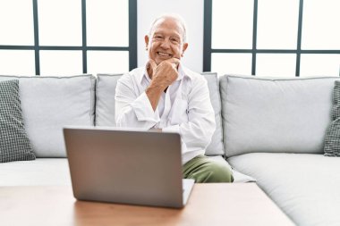 Senior man using laptop at home sitting on the sofa looking confident at the camera smiling with crossed arms and hand raised on chin. thinking positive. 