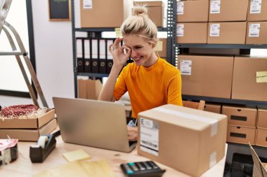 Young blonde woman working at small business ecommerce using laptop smiling happy doing ok sign with hand on eye looking through fingers 