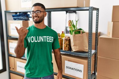 Young indian man volunteer holding donations box smiling with happy face looking and pointing to the side with thumb up. 