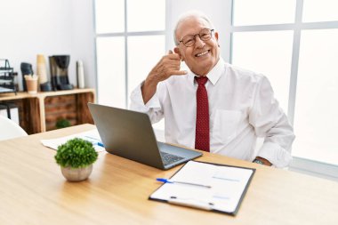 Senior man working at the office using computer laptop smiling doing phone gesture with hand and fingers like talking on the telephone. communicating concepts. 