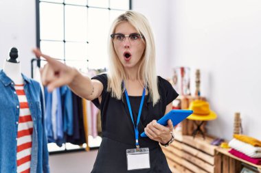 Young blonde woman working as manager at retail boutique pointing with finger surprised ahead, open mouth amazed expression, something on the front 