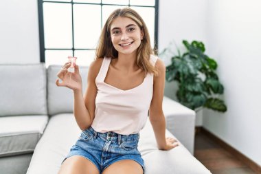 Elinde regl kupası tutan genç sarışın kadın pozitif ve mutlu görünüyor. Dişlerini gösteren kendine güvenen bir gülümsemeyle ayakta duruyor ve gülümsüyor. 