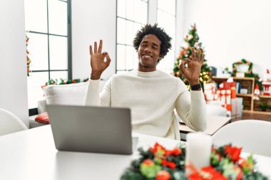 Genç Afro-Amerikalı adam dizüstü bilgisayar kullanıyor Noel ağacının yanında oturuyor rahat ve gözleri kapalı gülümsüyor parmaklarıyla meditasyon hareketi yapıyor. yoga konsepti. 