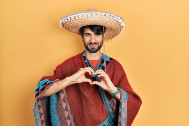 Young hispanic man holding mexican hat smiling in love doing heart symbol shape with hands. romantic concept. 