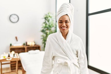 Havlu ve bornoz giymiş genç esmer kadın güzellik merkezinde mutlu ve havalı bir gülümsemeyle dikiliyor. şanslı kişi. 