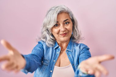 Middle age woman with grey hair standing over pink background smiling cheerful offering hands giving assistance and acceptance. 