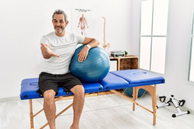 Orta yaşlı İspanyol adam ağrı kesici kliniğinde elinde pilates topu tutarken mutlu bir gülümsemeyle avuç içi yardım ve kabullenme sunuyor.. 