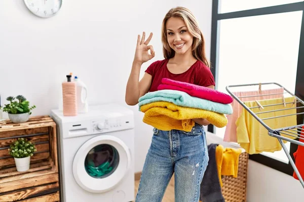 Young Caucasian Woman Holding Clean Laundry Doing Sign Fingers Smiling — Foto Stock