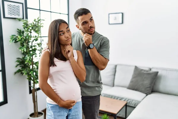 Young interracial couple expecting a baby, touching pregnant belly with hand on chin thinking about question, pensive expression. smiling with thoughtful face. doubt concept.