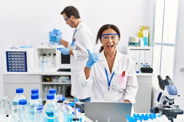 Middle age woman working at scientist laboratory pointing thumb up to the side smiling happy with open mouth