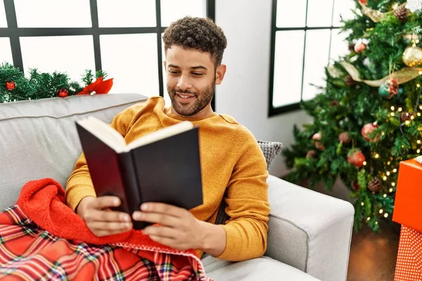 Young arab man reading book lying on the sofa by christmas tree at home.