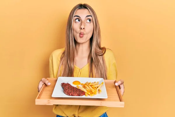 Beautiful hispanic woman holding tray with meat loaf and fried egg making fish face with mouth and squinting eyes, crazy and comical.