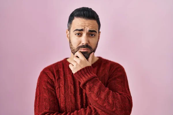 Young hispanic man with beard wearing casual sweater over pink background looking confident at the camera with smile with crossed arms and hand raised on chin. thinking positive.