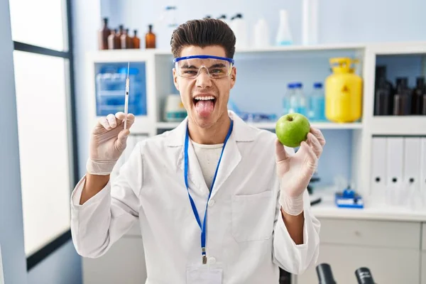 Young Hispanic Man Working Scientist Laboratory Holding Apple Sticking Tongue — ストック写真