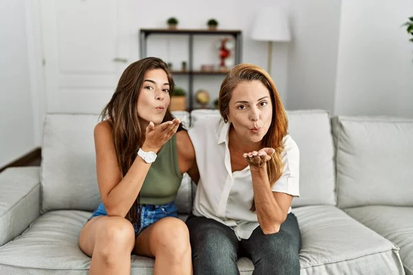 Mother and daughter together sitting on the sofa at home looking at the camera blowing a kiss with hand on air being lovely and sexy. love expression.