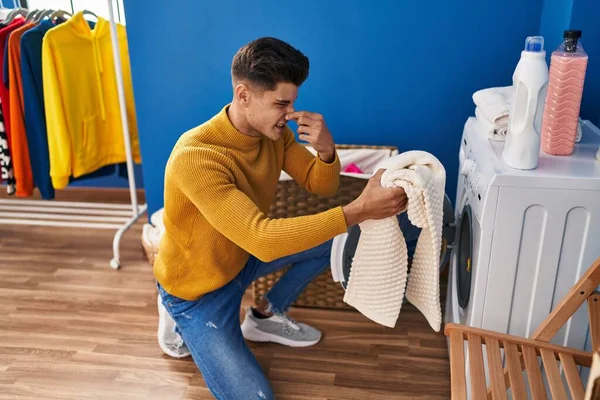 Young hispanic man washing clothes smelling dirty clothes at laundry