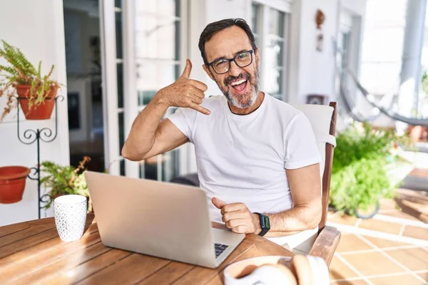 Middle age man using computer laptop at home smiling doing phone gesture with hand and fingers like talking on the telephone. communicating concepts.