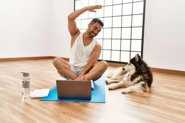 Young hispanic man smiling confident having online yoga class with dog at sport center