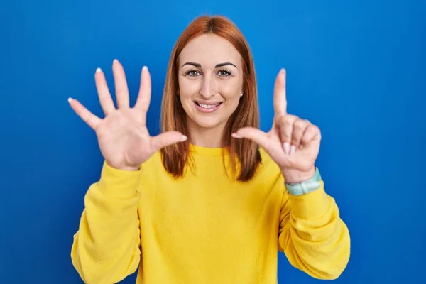 年轻的女人站在蓝色的背景上 用七号手指指指点点 自信而快乐地微笑着 — 图库照片