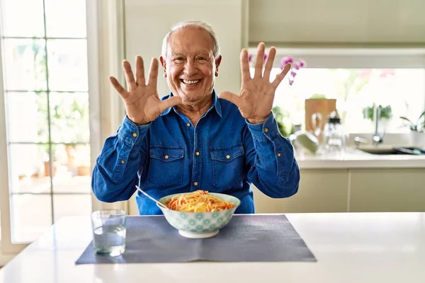 Senior man with grey hair eating pasta spaghetti at home showing and pointing up with fingers number ten while smiling confident and happy.
