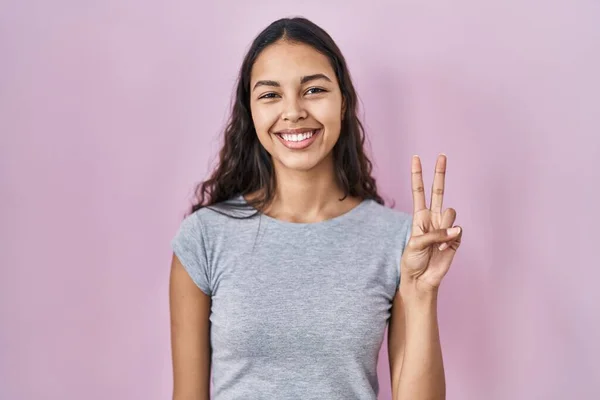 年轻的巴西女人穿着宽松的T恤 背景是粉色的 手指头指向二号 面带微笑 自信而快乐 — 图库照片