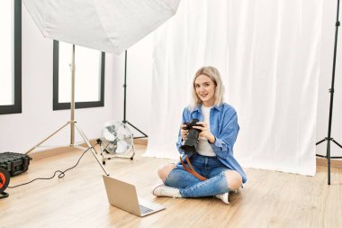 Fotoğrafçılık stüdyosunda çalışan güzel sarışın kadın yerde oturmuş bilgisayardaki fotoğrafları kontrol ediyor.