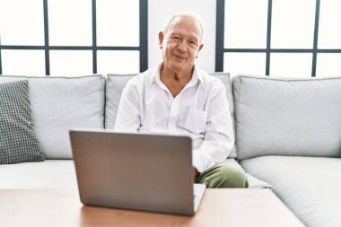 Senior man using laptop at home sitting on the sofa relaxed with serious expression on face. simple and natural looking at the camera. 