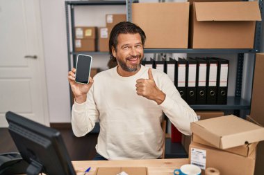Handsome middle age man working at small business ecommerce holding smartphone smiling happy and positive, thumb up doing excellent and approval sign 