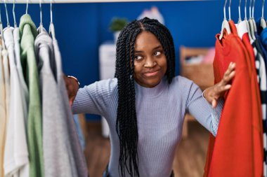 African american woman searching clothes on clothing rack smiling looking to the side and staring away thinking. 