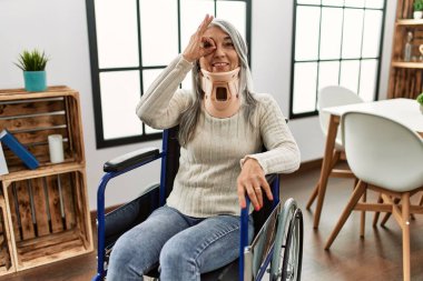Middle age grey-haired woman sitting on wheelchair with collar at home smiling happy doing ok sign with hand on eye looking through fingers 