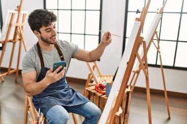 Genç İspanyol ressam mutlu bir resim çiziyor ve resim stüdyosunda akıllı telefon kullanıyor..