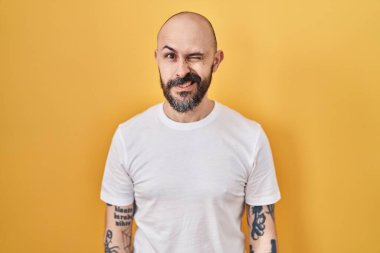 Young hispanic man with tattoos standing over yellow background winking looking at the camera with sexy expression, cheerful and happy face. 
