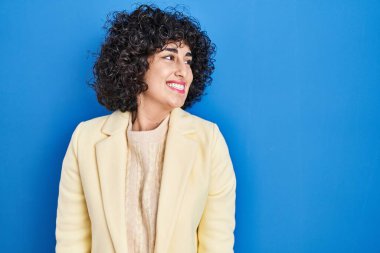 Young brunette woman with curly hair standing over blue background looking away to side with smile on face, natural expression. laughing confident. 