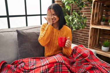 Kanepede oturan genç İspanyol kadın evde kahve içiyor bir gözünü eliyle kapatıyor, yüzünde kendine güvenen bir gülümseme ve sürpriz bir duygu var.. 