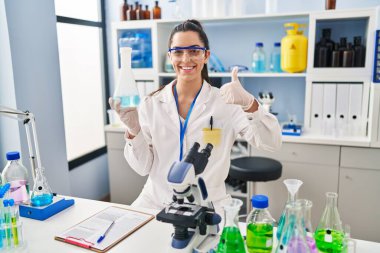 Hispanic woman working at scientist laboratory smiling happy and positive, thumb up doing excellent and approval sign 