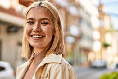 Genç sarışın kız gülümsüyor, mutlu bir şekilde şehirde duruyor..