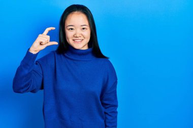 Young chinese girl wearing casual clothes smiling and confident gesturing with hand doing small size sign with fingers looking and the camera. measure concept. 
