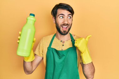 Young hispanic man wearing cleaner apron holding cleaning product pointing thumb up to the side smiling happy with open mouth 