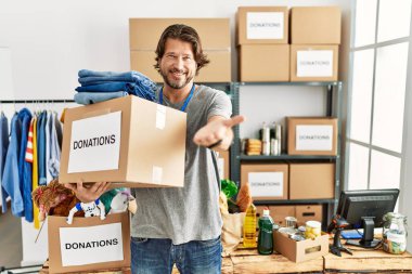 Handsome middle age man holding donations box for charity at volunteer stand smiling cheerful offering palm hand giving assistance and acceptance. 