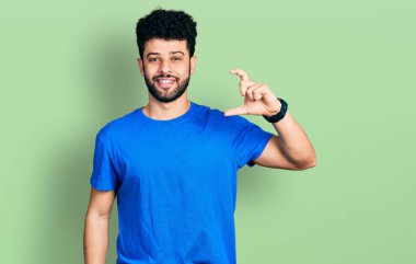Sakallı, günlük mavi tişörtlü, gülümseyen, kendine güvenen, el hareketi yapan, parmaklarıyla işaret yapan ve kameraya bakan genç bir Arap. ölçüm kavramı. 