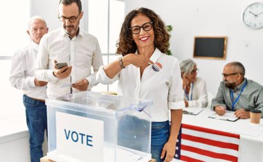 Middle age american voter woman smiling happy pointing with finger to usa badge at vote center.